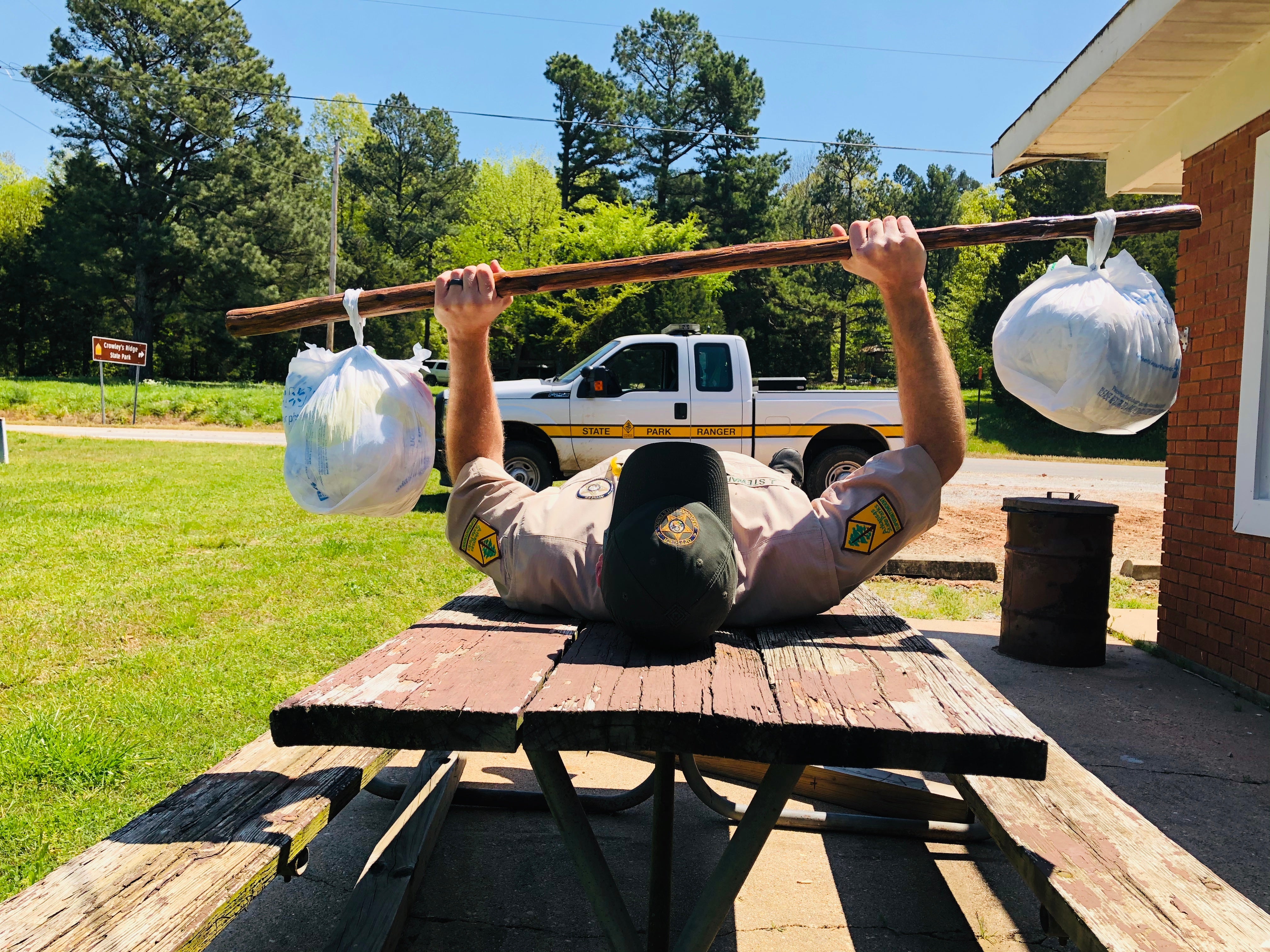 Park ranger lying on his back on top of a picnic table, lifting plastic bags tied to the ends of a hiking stick like one would lift weights 